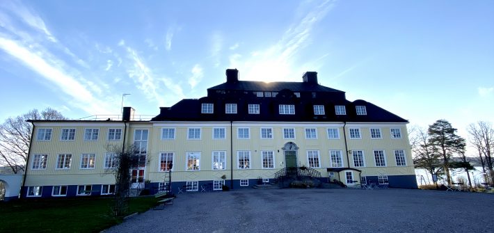 Bilden visar Rimforsa strand. En gul herrgårdsliknande hotellbyggnad i Rimforsa vid sjön Åsunden i Östergötland